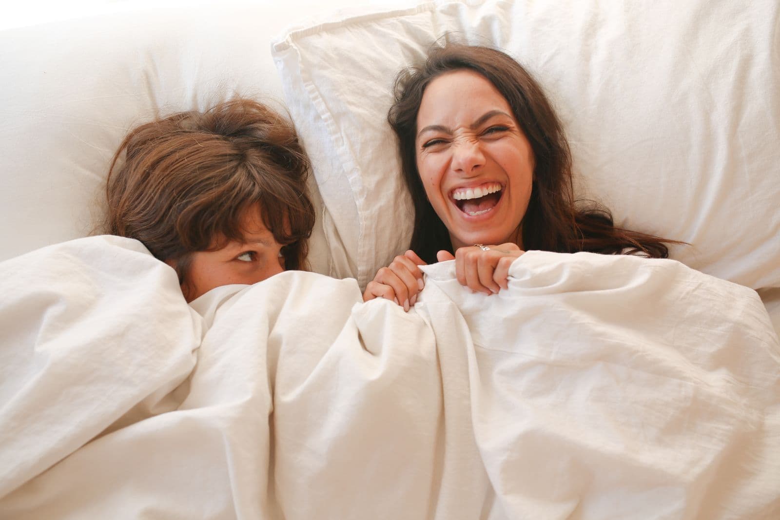 Two women lying in bed under white sheets, one laughing. Photo by RDNE Stock project on Pexels.