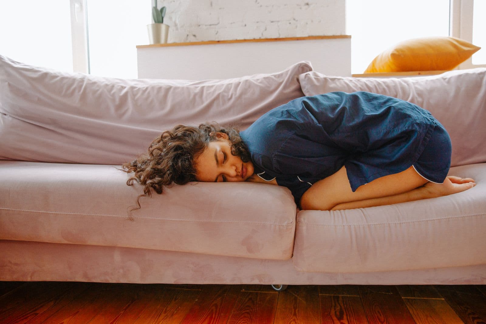 A woman curled up asleep on a sofa in daylight. Photo by Polina Zimmerman on Pexels.