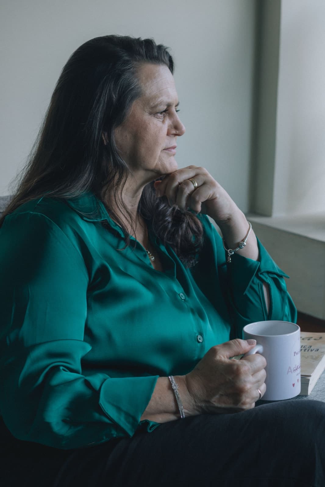 Middle-aged woman holding a white mug beside a softly lit window. Photo by Mayara Caroline Mombelli on Pexels.