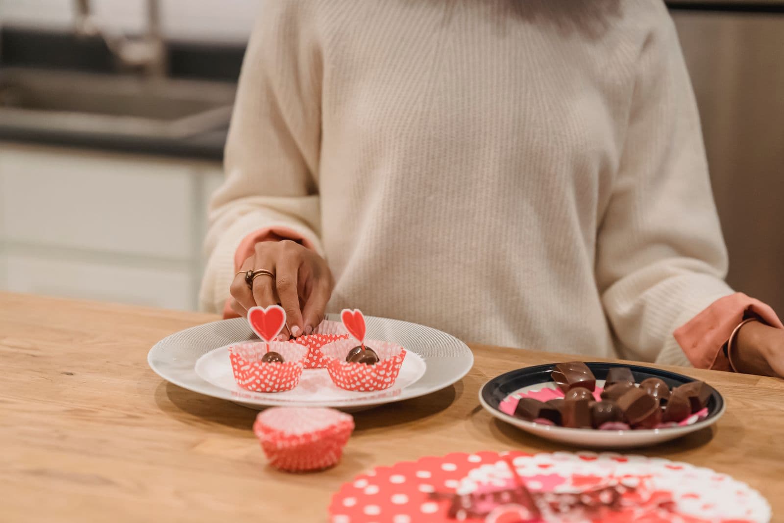 A woman in a cream jumper at a kitchen counter with a plate of chocolates in front of her. Photo by Andres Ayrton on Pexels.