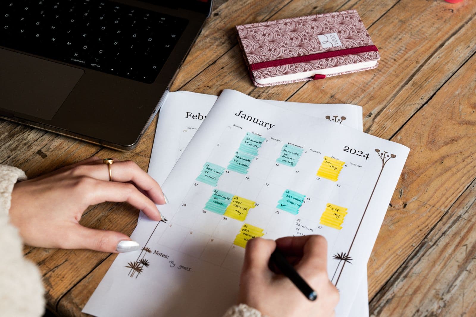Hands marking dates on a paper calendar with a pen, beside a laptop and a notebook. Photo by Ahmed on Pexels.