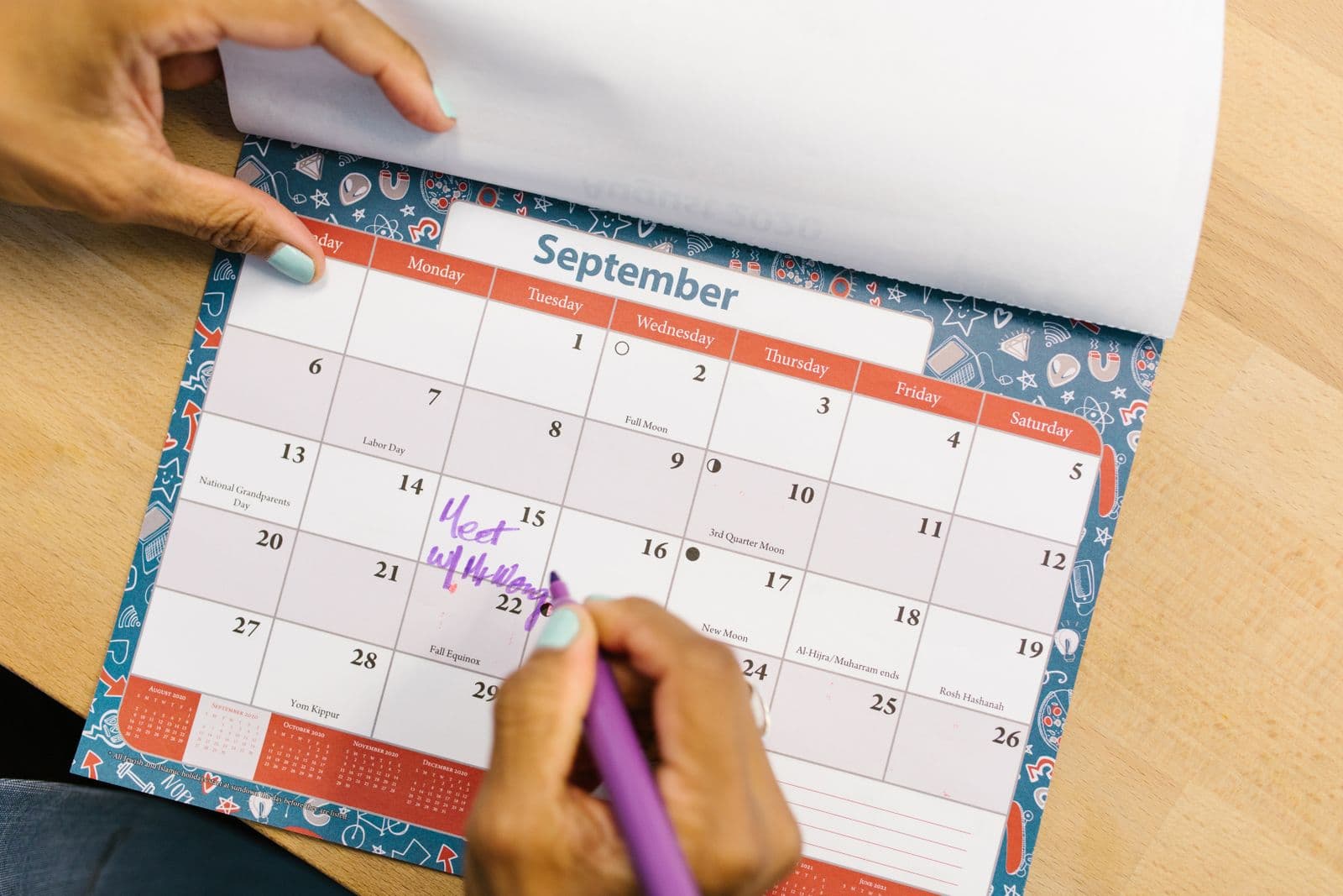 A person marking a date on a paper wall calendar with a purple pen on a wooden desk
