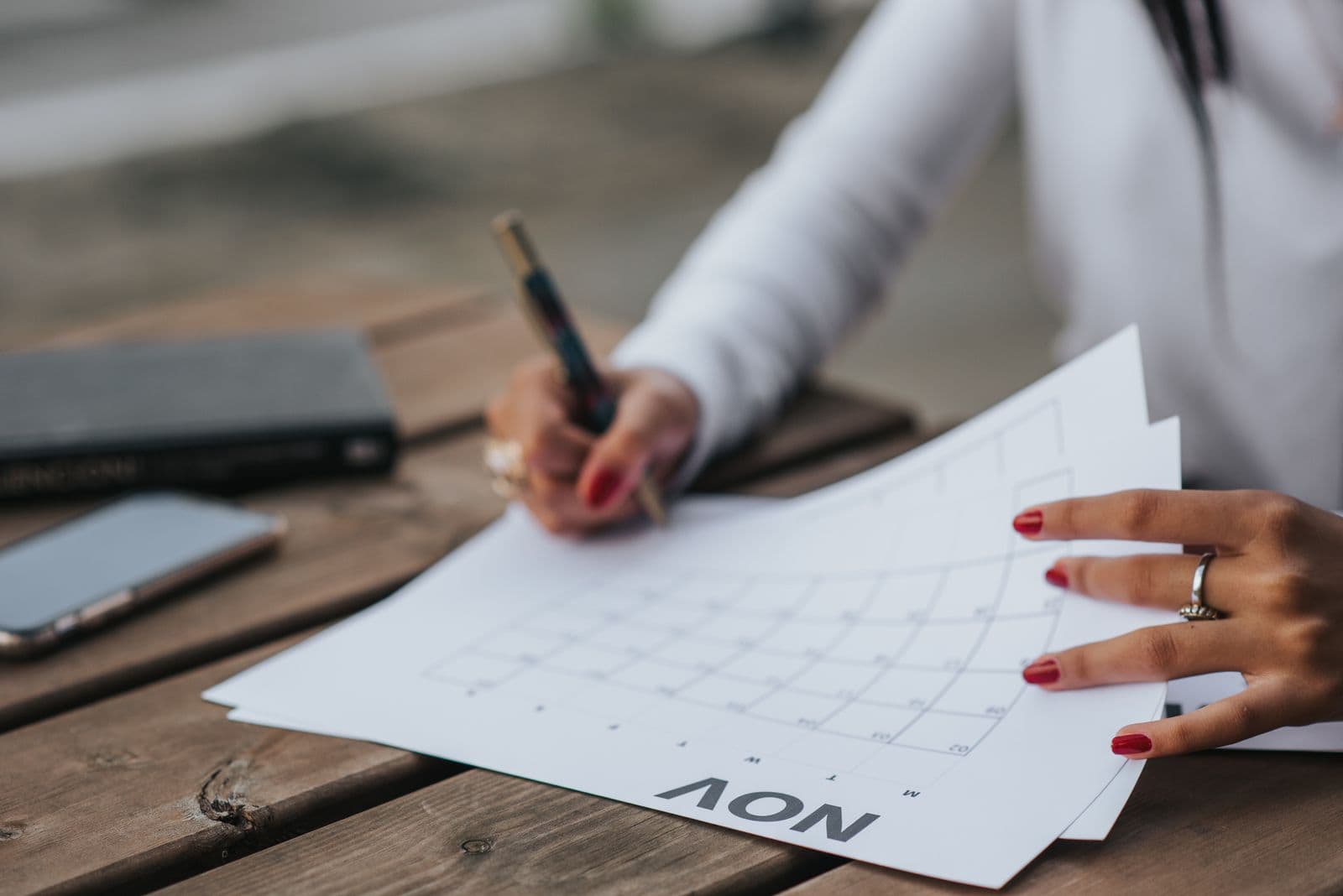 A woman sitting outdoors at a wooden table, writing in a paper calendar. Photo by Anete Lusina on Pexels.