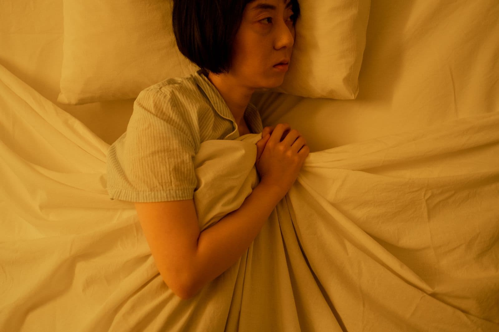 A woman with short dark hair lying awake in bed at night, holding the sheet to her chest in warm, dim light. Photo by cottonbro studio on Pexels.