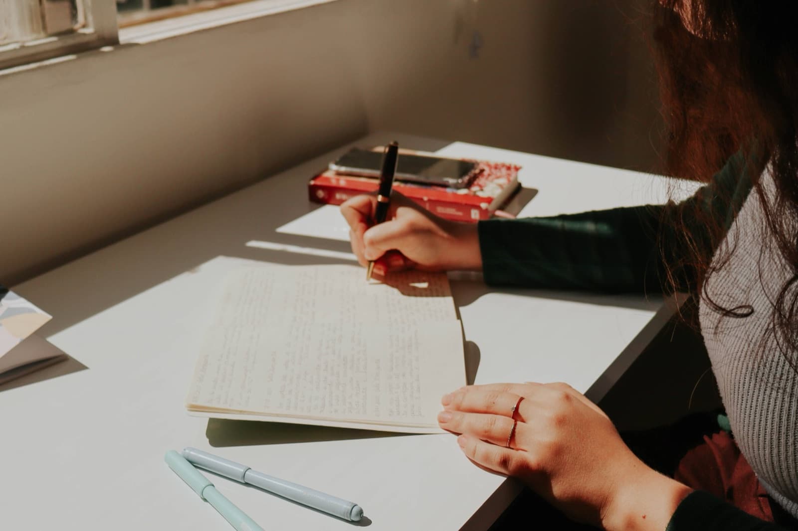 A woman writing in a notebook beside a sunlit window. Photo by Letícia Alvares on Pexels.