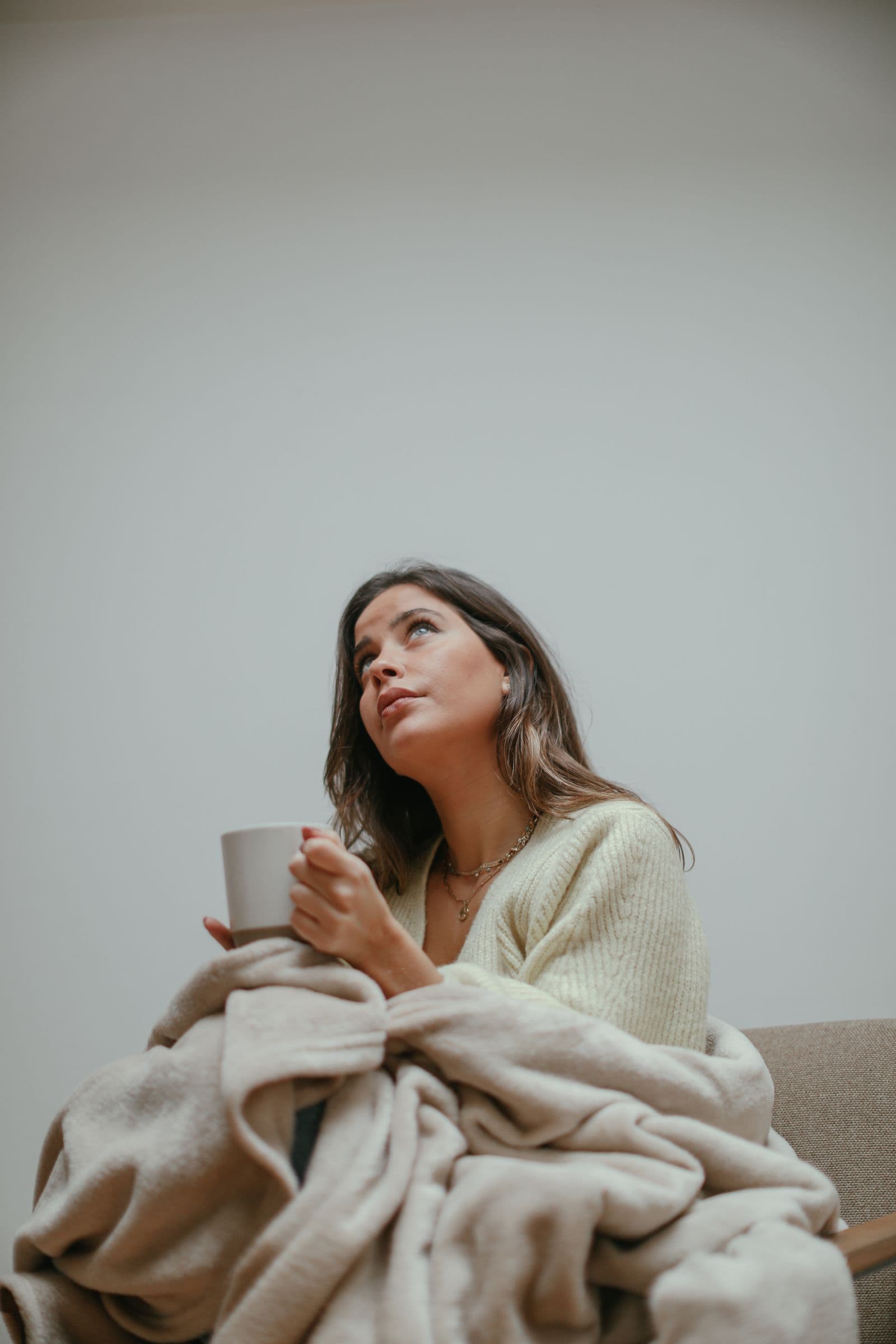 A woman wrapped in a cream blanket holding a warm mug in soft indoor light. Photo by Cup of Couple on Pexels.