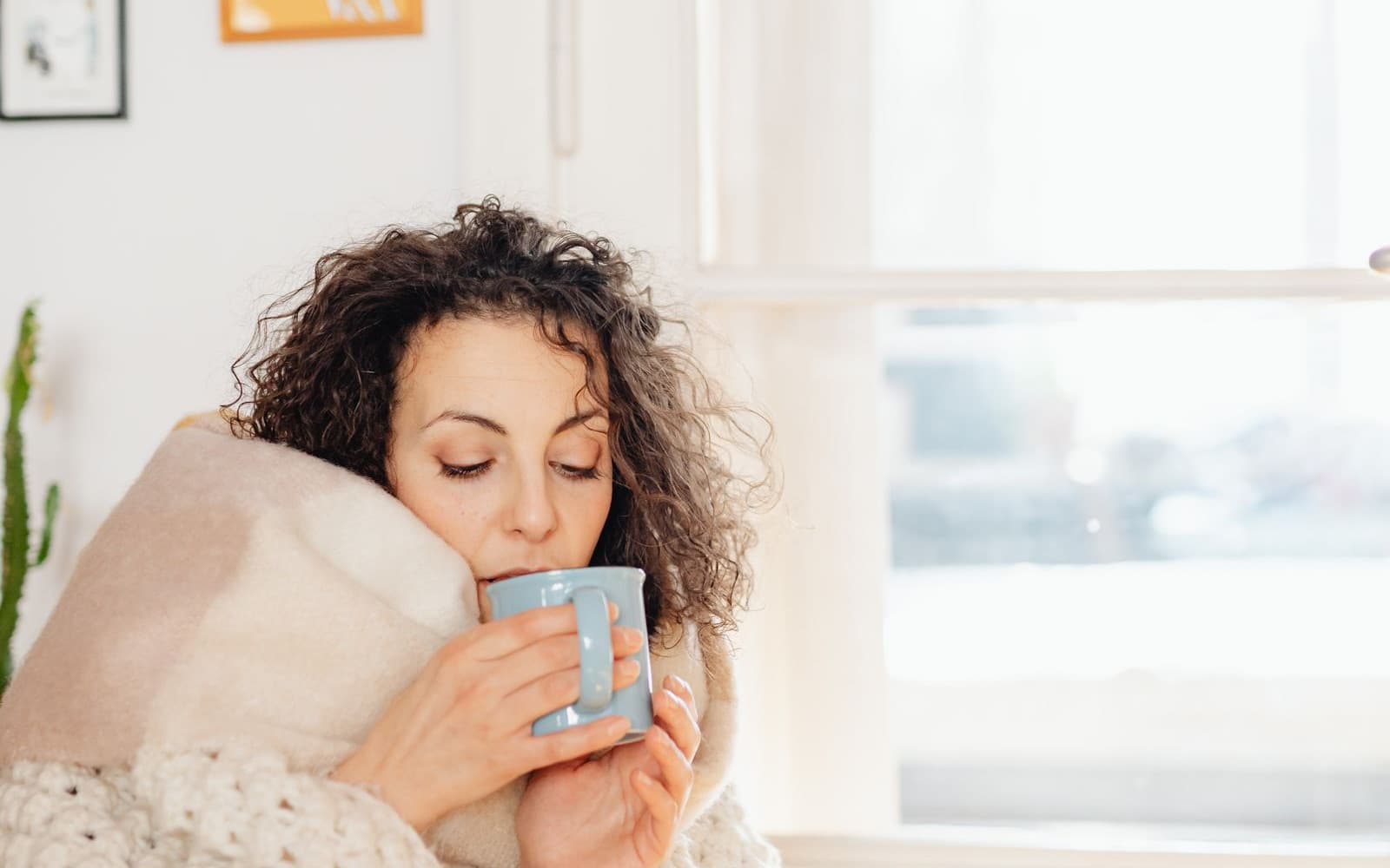 A woman wrapped in a soft blanket holding a warm mug beside a bright window. Photo by Kaboompics.com on Pexels.