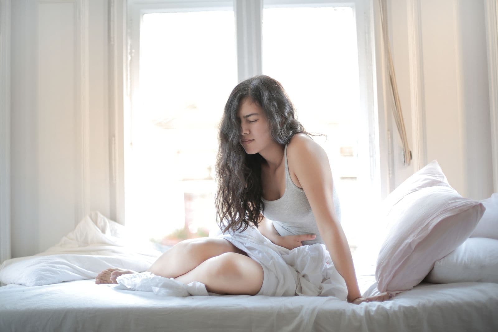 A woman sitting on a bed with both hands pressed to her lower abdomen. Photo by Andrea Piacquadio on Pexels.