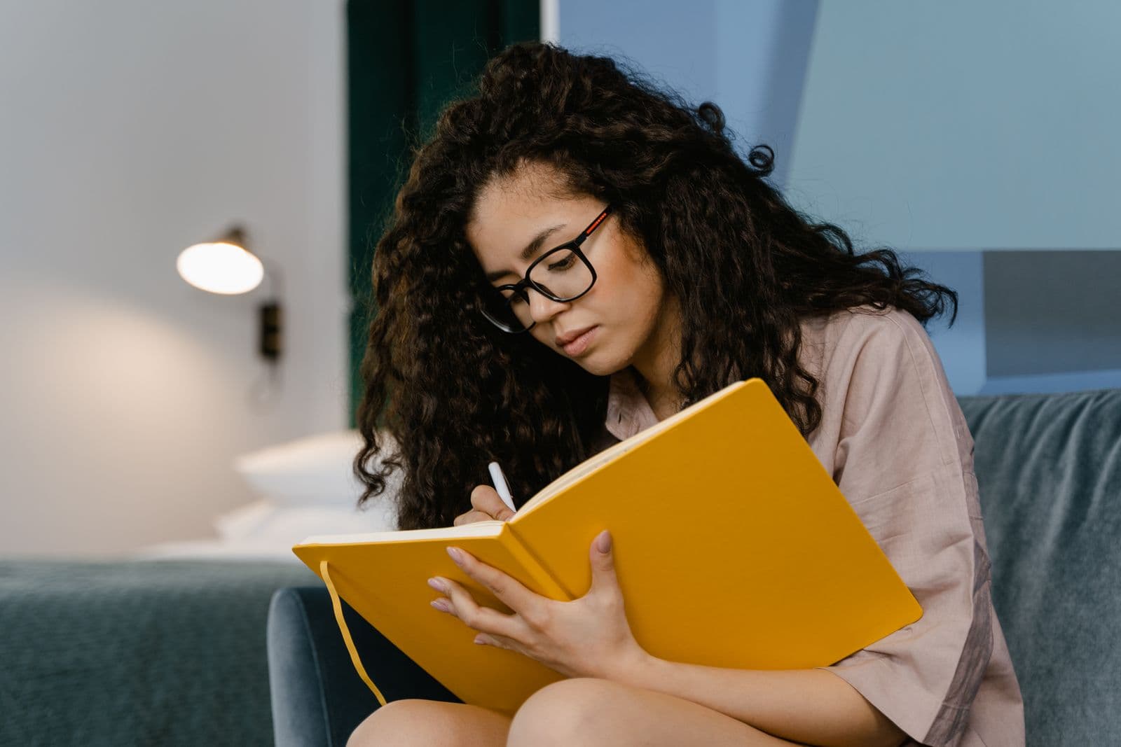 A young woman with curly hair writing in a yellow notebook at a table. Photo by MART PRODUCTION on Pexels.