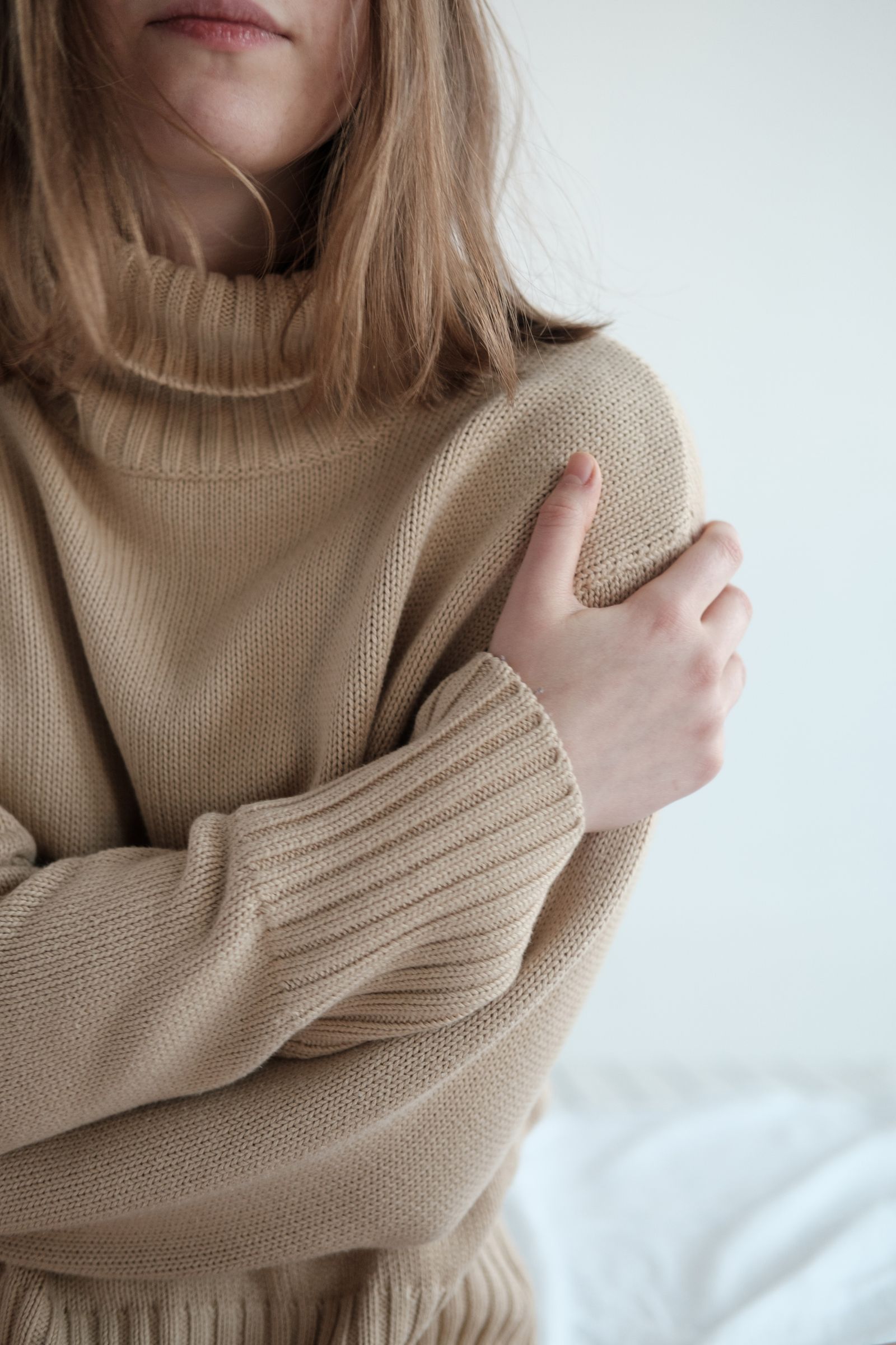 A person in a warm sweater sitting on a bed with arms folded