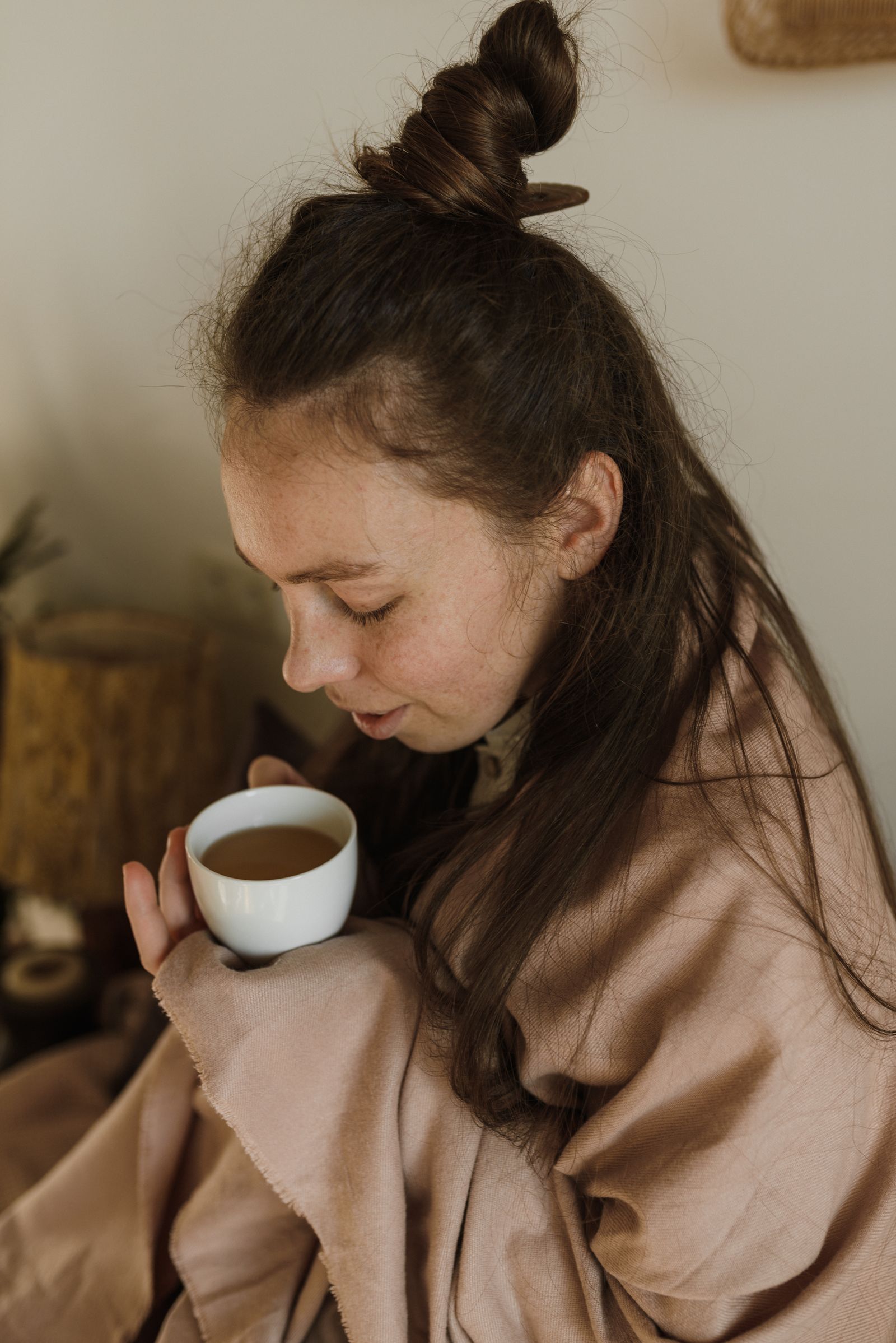 Woman wrapped in a soft neutral blanket holding a warm drink at home