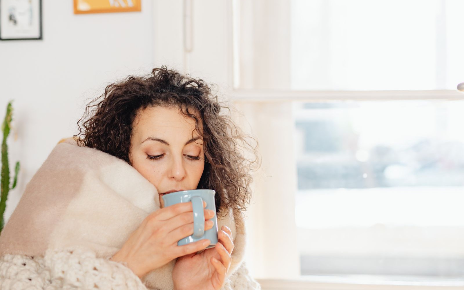 A woman wrapped in a soft blanket holding a warm mug beside a bright window
